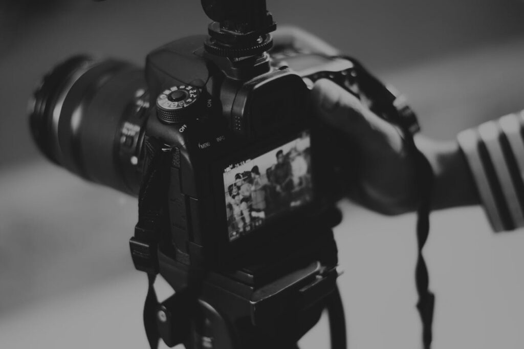 The black-and-white image captures a close-up view of a person holding a DSLR camera, focusing on the camera's rear screen. The screen displays a preview of a group photo being taken. The camera has various control dials and buttons visible, with the large lens extending outward. The person's hand, partly out of focus, is adjusting the camera, which is mounted on a tripod or stabilizer.