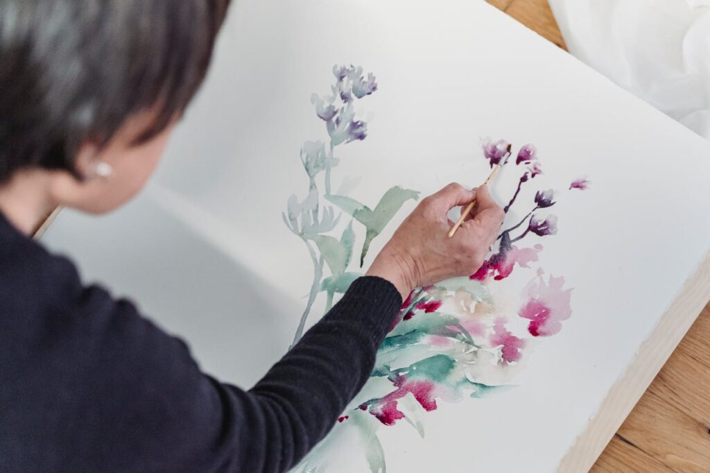 Close-up image of a woman painting delicate floral designs on a large sheet of paper. The artwork appears to be in a watercolor style, featuring soft greens, reds, and purples. The individual is holding a brush and focusing on the details of the flowers. The person is wearing a dark long-sleeved shirt, and the painting area is well-lit, possibly near a window or under natural light. The floral patterns suggest a gentle, flowing style of painting, with soft color transitions and a serene composition. Representing Craft hobbies for moms in their 40s.