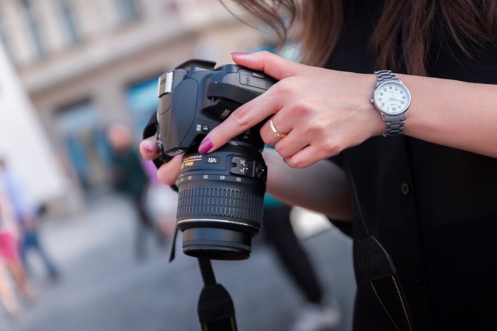 A close-up shot of a woman holding a DSLR camera, adjusting its settings with her right hand. She is wearing a silver wristwatch and has neatly manicured nails painted pink. The camera lens is detailed, with focus and zoom rings visible, suggesting she is preparing for a shot. The background is softly blurred, showing an urban outdoor setting with a few indistinct people and buildings, giving the impression of street photography.