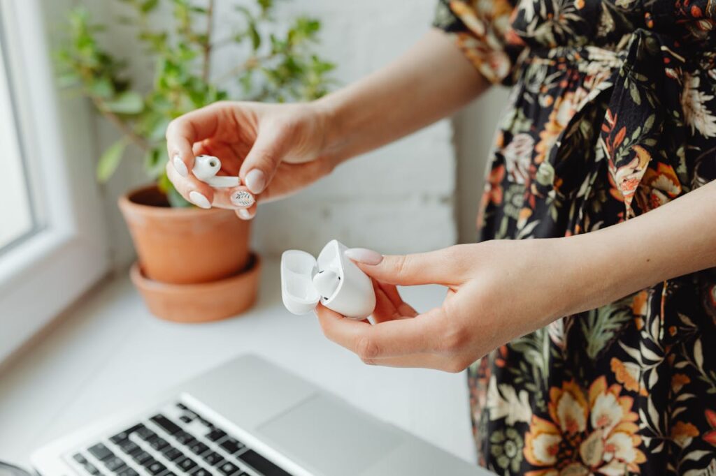 Image of a woman holding a pair of white wireless earbuds and their charging case, preparing to place one earbud into the case. They are standing near a desk with an open laptop in the foreground. The individual is wearing a dark floral dress, and in the background, there are small potted plants on a windowsill, adding a touch of greenery to the scene. The natural lighting from the window enhances the peaceful and productive atmosphere, suggesting a moment of technology use in a cozy home environment.