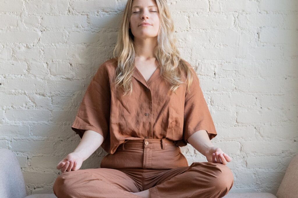 Image of a women seated cross-legged on a soft surface, meditating with their eyes closed, exuding a sense of calm and tranquility. They are wearing a loose-fitting, earthy-toned outfit in shades of brown. Their hands rest gently on their knees with palms facing upward in a traditional meditation posture. The background is a light-colored brick wall, which gives the scene a minimalist and peaceful ambiance. The soft lighting enhances the relaxed, mindful atmosphere.