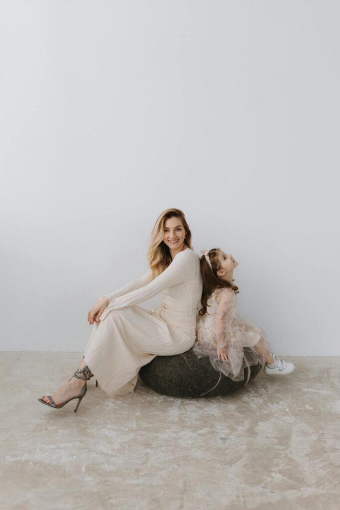 Image of a woman and a young girl sitting back-to-back on a large, round cushion. The woman is smiling, dressed in a flowing, cream-colored dress with high heels, while the little girl is wearing a light, frilly dress with a headband and sneakers. The little girl is looking up with a playful expression. The background is minimal, with soft, neutral tones, giving the image a light and elegant feel, highlighting the gentle connection between the two.