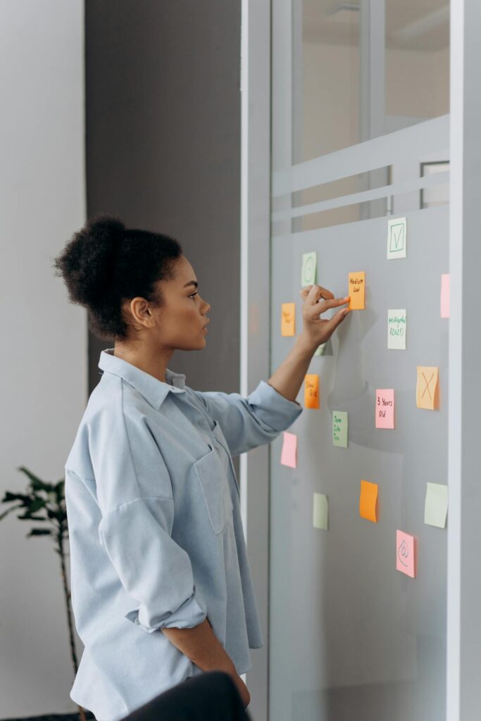 Image of a woman standing next to a glass wall, organizing colorful sticky notes. She is wearing a light blue shirt with rolled-up sleeves and is seen placing one of the notes on the wall. The notes come in various colors, including orange, pink, green, and yellow, and appear to contain different types of tasks or ideas. The scene suggests a brainstorming or planning session in an office environment. In the background, there is a small plant, adding to the professional yet relaxed atmosphere.