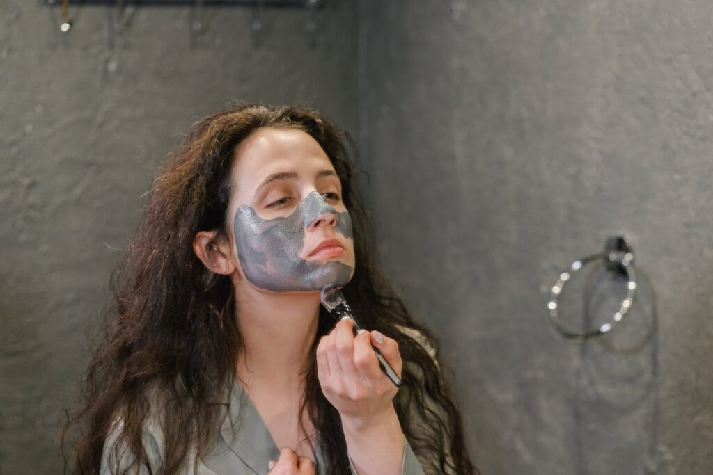 Image of a woman applying a gray facial mask on her face using a brush. she appears to be in a bathroom with dark, minimalist background. her long dark hair is loose and se looks focused on the application.