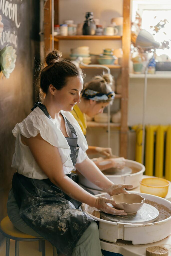 Image of two women engaged in pottery making in a cozy, sunlit studio. The woman in the foreground, wearing a white blouse and a black apron, shapes clay on a spinning pottery wheel with a focused expression. In the background, another woman, wearing a headband and a yellow top, also works on her pottery. Shelves filled with ceramic pieces and pottery tools can be seen in the background, creating a creative and artistic atmosphere.