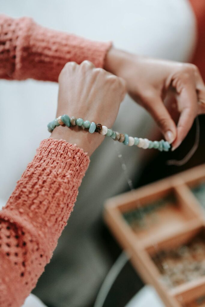 Image of a woman threading beads onto a string, with hands visible holding the strand. The beads are of various shades of blue, green, and beige. The person is wearing a textured, long-sleeve coral-colored sweater. A wooden tray with organized compartments is slightly blurred in the background.