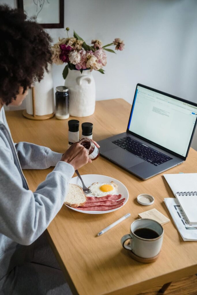 Image of a person is sitting at a wooden table, having breakfast while working on a laptop. The meal consists of a plate with fried eggs, strips of bacon, and slices of bread. The person is spreading a condiment, possibly jam or jelly, onto the bread. A cup of coffee sits next to an open notebook and a pen. In the background, a vase with pink and white flowers adds a touch of decor. The laptop screen shows a document, suggesting the person is multitasking, possibly working from home.