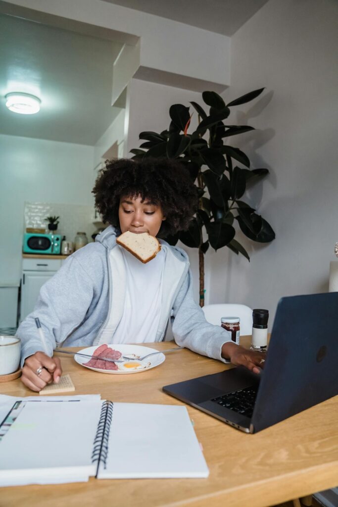 Close-up image of a woman multitasking at a table, eating breakfast while working. They have a slice of bread held in their mouth, a plate of food (including eggs and sausage) on the table, and they are using one hand to write in a notebook and the other to work on a laptop. They are wearing a light gray hoodie, and in the background, there is a kitchen area and a large potted plant. The scene suggests a busy morning, combining work and breakfast representing  be a successful stay at home mom.