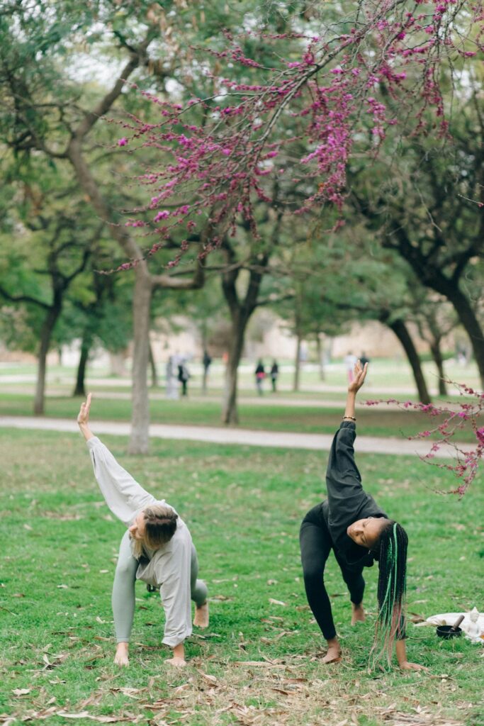 Image of two people are practicing yoga outdoors on a grassy field under trees with blooming pink flowers. They are in a side stretch pose, each bending to the side with one arm extended upward and the other reaching down toward the ground. The scene feels calm and serene, with the surrounding trees and open space adding to the peaceful atmosphere. Both individuals are dressed in comfortable, loose-fitting clothes.