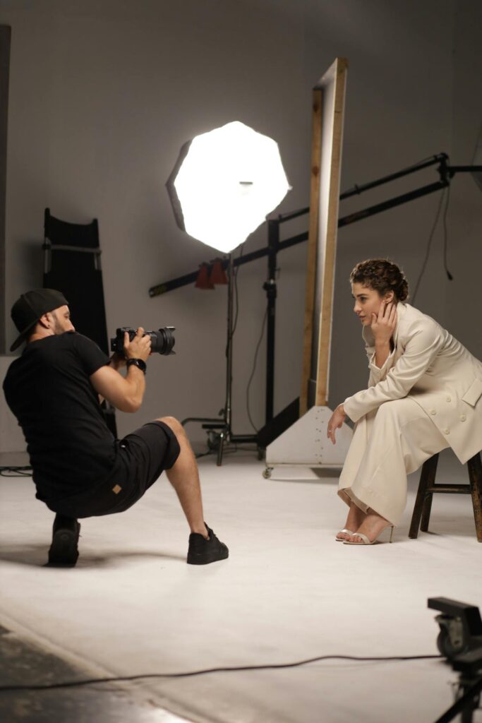 Image of studio setup with a photographer,  crouching and taking a photo of a model, whose face is also obscured. The model is seated on a white block wearing a light-colored suit. The studio has various equipment including lighting, reflectors, and stands. The background is neutral and unfocused.