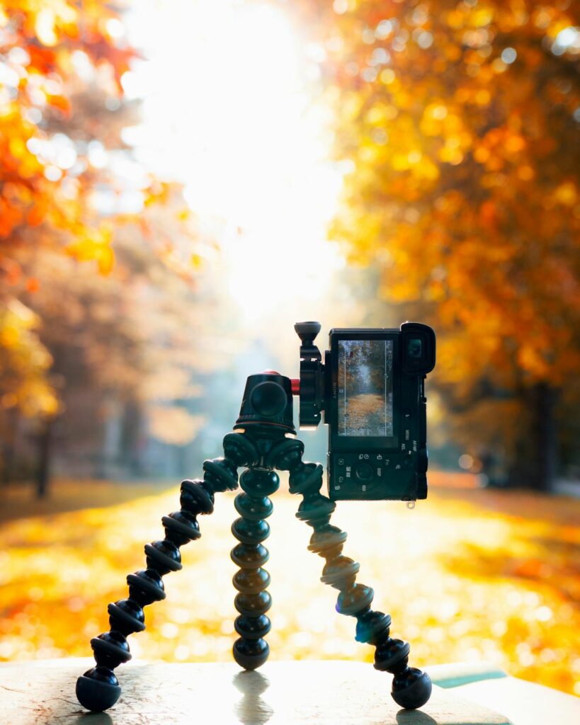 Image of a camera mounted on a flexible tripod is positioned outdoors, capturing an image of a sunlit autumn scene with trees displaying vibrant orange and yellow leaves. The camera’s screen shows the colorful foliage bathed in the bright sunlight, creating a warm and picturesque atmosphere. The focus on the camera setup against the blurred background highlights the act of photography amidst the beauty of fall.