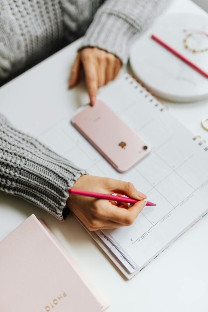 Close-up image of a person is sitting at a desk, writing in a planner or calendar with a pink pencil. They are wearing a cozy gray sweater, and their other hand is pointing to a date on the calendar. A pink smartphone lies on the desk next to the planner, and nearby are a closed pink notebook and a white round tray with a pencil and small jewelry. The scene suggests someone organizing their schedule or making plans. The soft lighting and coordinated pastel colors create a calm, productive atmosphere.