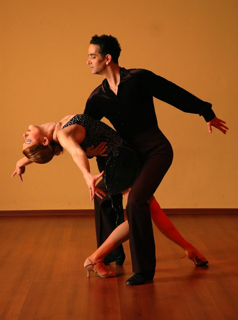 Image of a man and a woman perform a dance in an elegant pose. The man, dressed in dark clothing, supports the woman as she leans backward with an arched back and extended arms, wearing a sleeveless black dress and heels. They both appear focused, with graceful body positioning, as they dance on a polished wooden floor against a plain, warm-toned background. Image representing the dance hobbies.