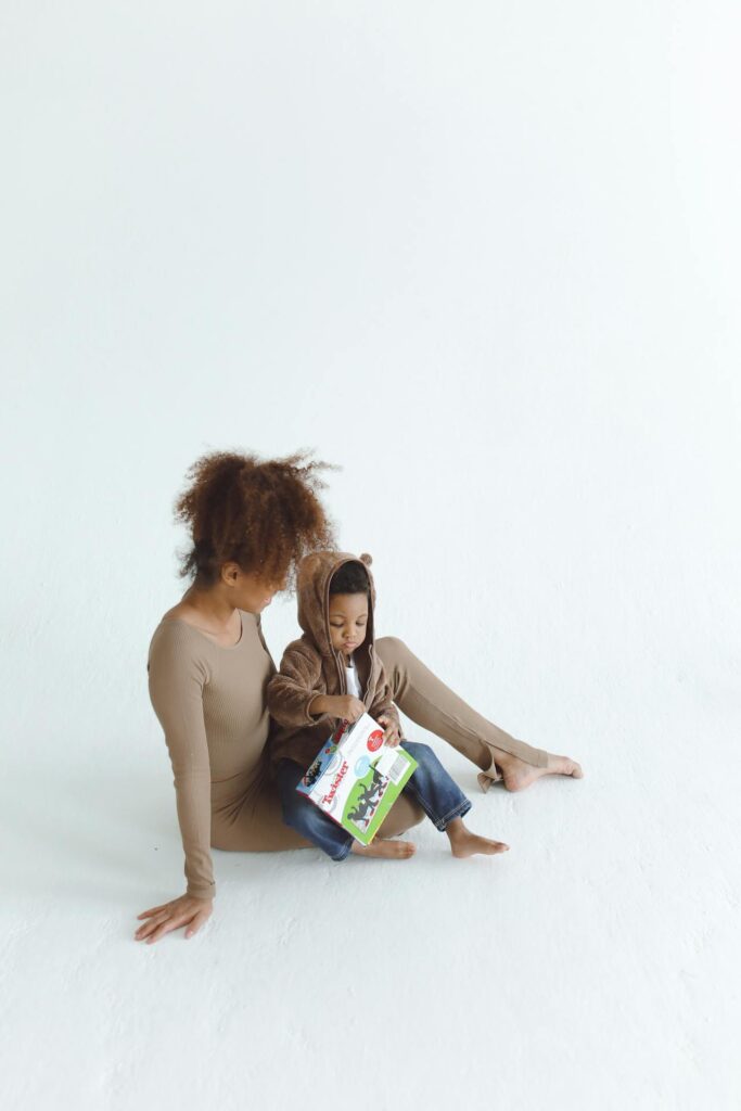 Image of a young woman sitting on the floor next to a small child, who is engrossed in a picture book. The woman is dressed in a long, beige bodysuit, while the child is wearing a cute hoodie with bear ears. They are both barefoot, sitting on a bright white floor against a plain white background, creating a minimalist and serene atmosphere. 