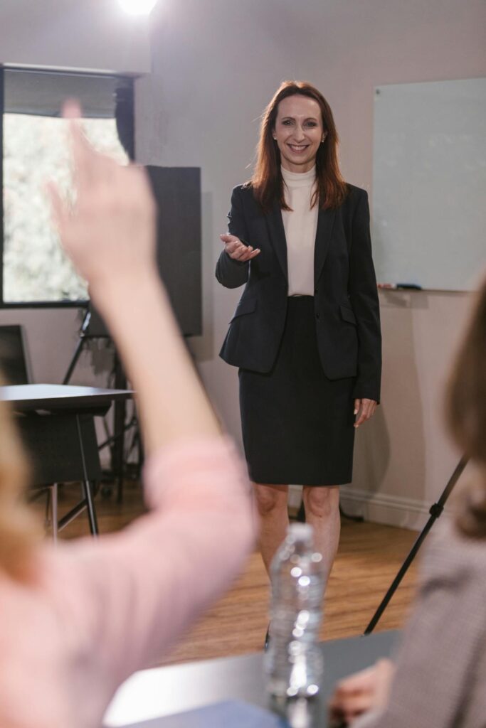 Image of a professional woman standing at the front of a room, likely leading a presentation or classroom session. She is wearing a formal dark blazer and skirt with a light-colored turtleneck, smiling confidently as she gestures toward someone in the audience. In the foreground, a participant is raising their hand, and a bottle of water is on the table. The setting suggests an interactive session, with the woman engaging her audience in a professional, educational, or corporate environment.
