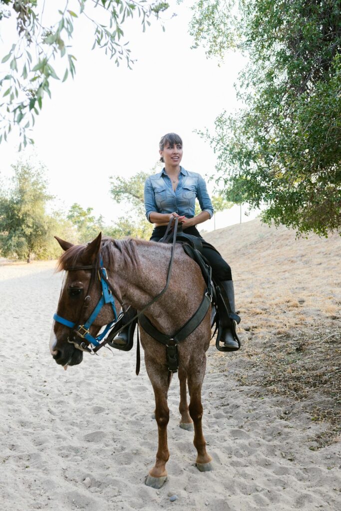 Image of a woman riding a horse along a sandy path in a natural, outdoor setting. She wears a blue denim shirt and black pants, smiling as she holds the reins. The horse, with a brownish-gray coat and a blue bridle, stands calmly beneath the shade of nearby trees. The background includes greenery and dry grass, suggesting a peaceful trail ride showing boredom in your 40s.