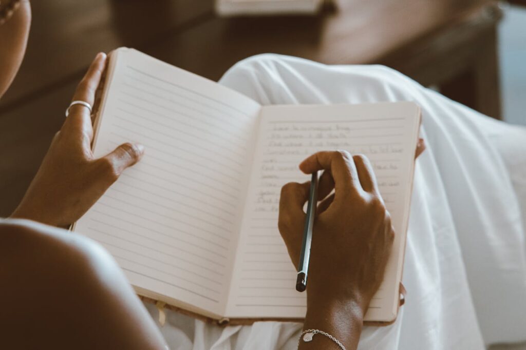 Close-up image of a person is sitting with a notebook open on their lap, writing with a pen on lined pages. They wear a ring and bracelet, and their relaxed posture suggests a calm, reflective moment. The warm lighting and blurred background create a cozy, intimate atmosphere focused on the act of journaling or writing.