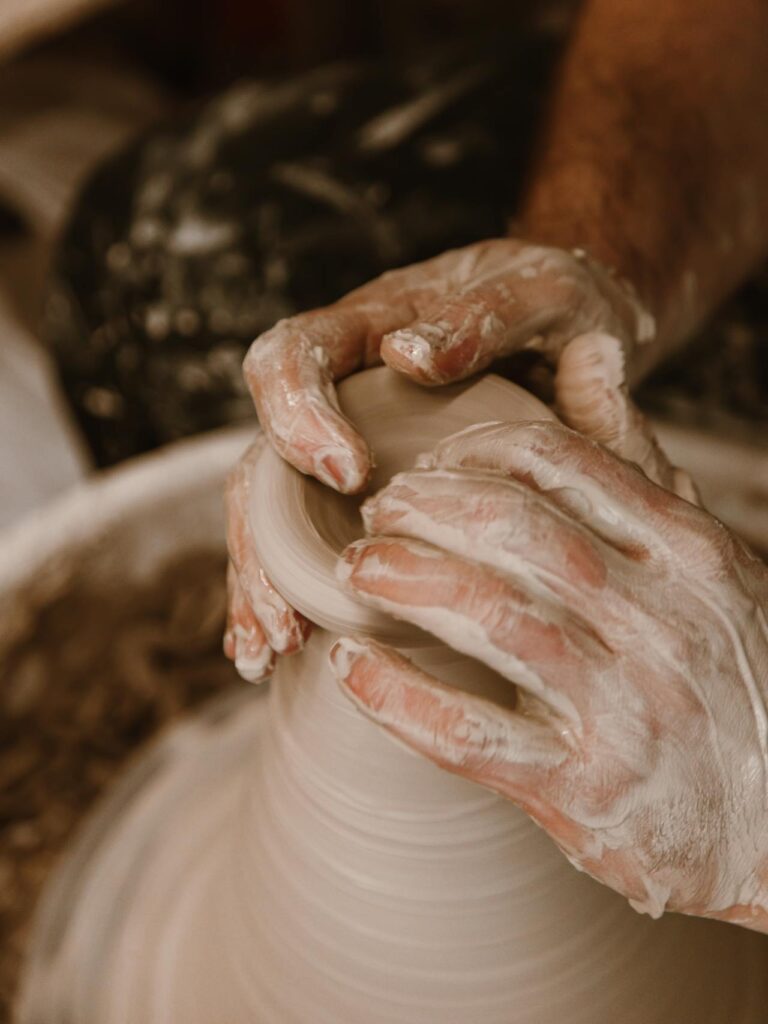 A close-up image of a person's hands shaping clay on a pottery wheel. The hands are covered in wet clay, and the spinning motion of the wheel creates a blurred effect on the clay as it is molded into a cylindrical form showing Cute hobbies for women.