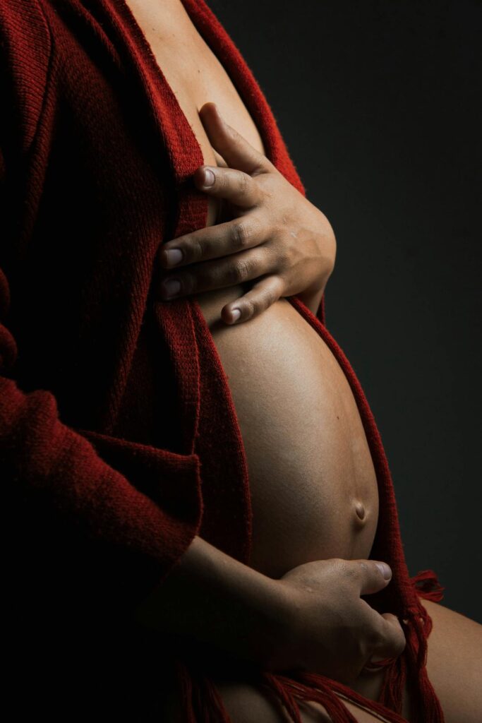 Close-up image of a pregnant belly, with a person gently cradling it using both hands. The individual is wearing a dark red robe, partially open to reveal the belly. The lighting is soft and dramatic, creating shadows that emphasize the curves of the body and the warmth of the skin. The overall mood of the image feels intimate and serene.