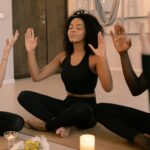 Image of group of people sitting on yoga mats in a relaxed meditation session. A woman in the center, with curly hair, wearing black workout clothes, sits cross-legged with her eyes closed and hands raised, as if focusing on deep breathing. Candles and flowers are placed on the floor, creating a calm and serene atmosphere. Image used for the article Hobbies for Moms in Their 40s.