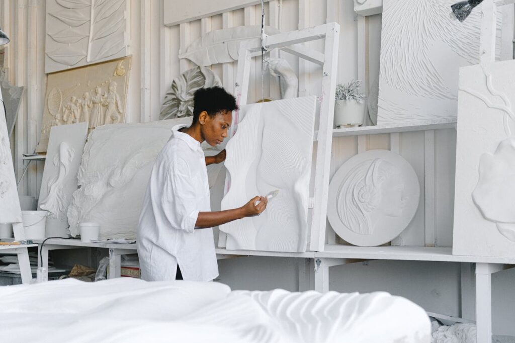 Image of an artist, dressed in a loose white shirt, works intently on a large white relief sculpture in a studio filled with similar artworks. They are holding a tool and carefully shaping intricate textures on the surface. The studio has shelves and walls displaying various textured and embossed art pieces, all in shades of white, creating a calm, monochromatic environment that emphasizes the details of the sculptures.