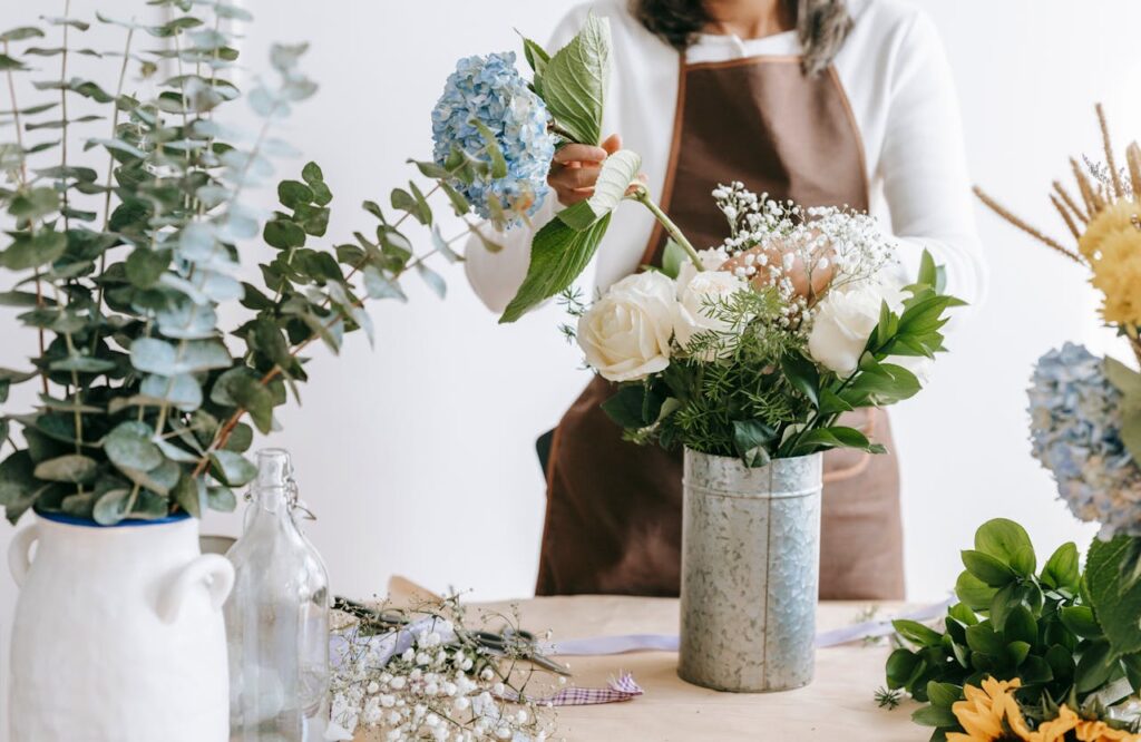 Close-up image of a woman arranging a flower bouquet at a table, with a variety of flowers and greenery. The arrangement includes white roses, blue hydrangeas, and baby's breath in a rustic metal vase. The person is wearing a white shirt and brown apron, with other floral arrangements and tools visible around the table.