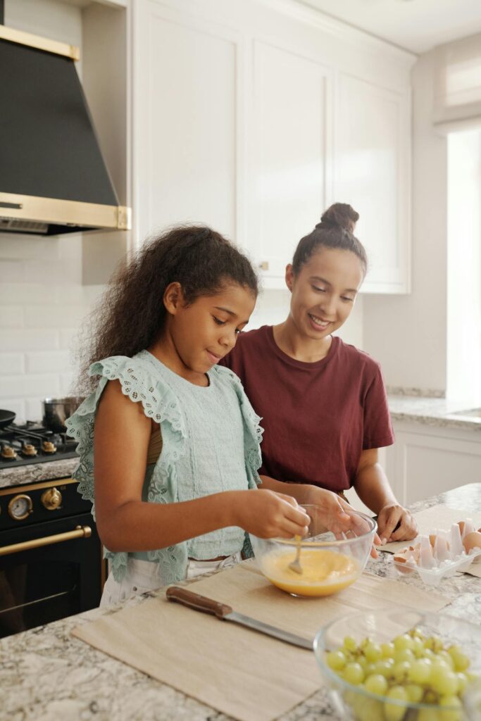 Image of two girls are in a bright, modern kitchen, preparing food together. The girl on the left, wearing a light blue sleeveless top, is whisking eggs in a glass bowl, while the girl on the right, in a maroon shirt, smiles as she watches. On the counter, there are cracked eggshells, a knife, and a bowl of green grapes. The scene captures a happy, collaborative moment as the two girls enjoy cooking together.