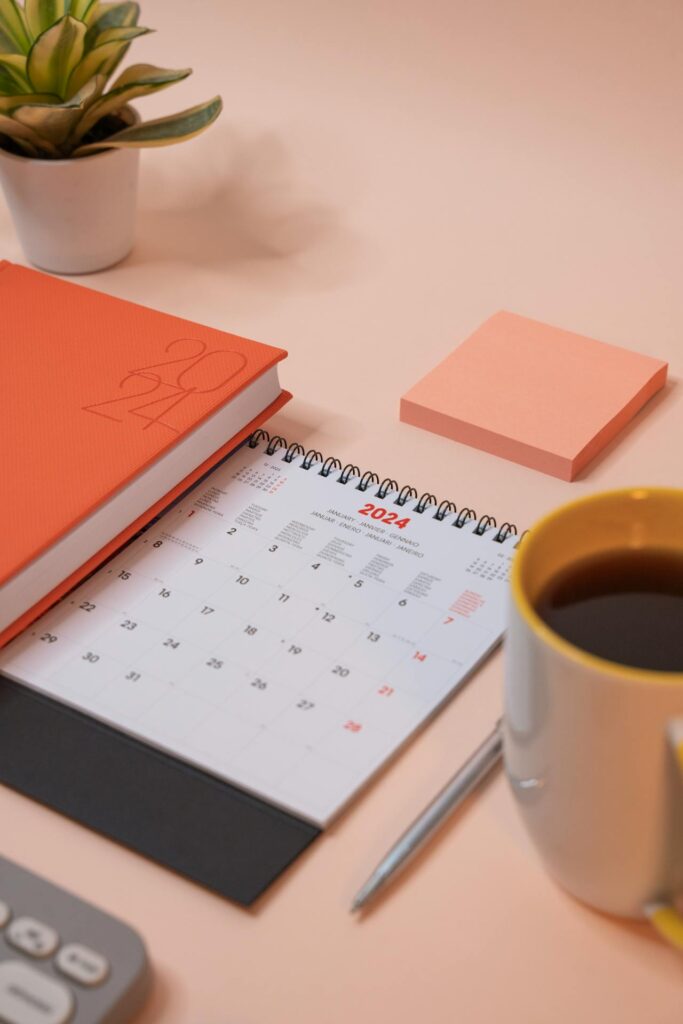 Image of a desk calendar is placed on a light-colored surface alongside an orange notebook, a silver pen, a peach-colored sticky note pad, a coffee mug, and a small potted plant. The scene has an organized and minimalist feel, with a calm color scheme centered around peach, orange, and neutral tones.