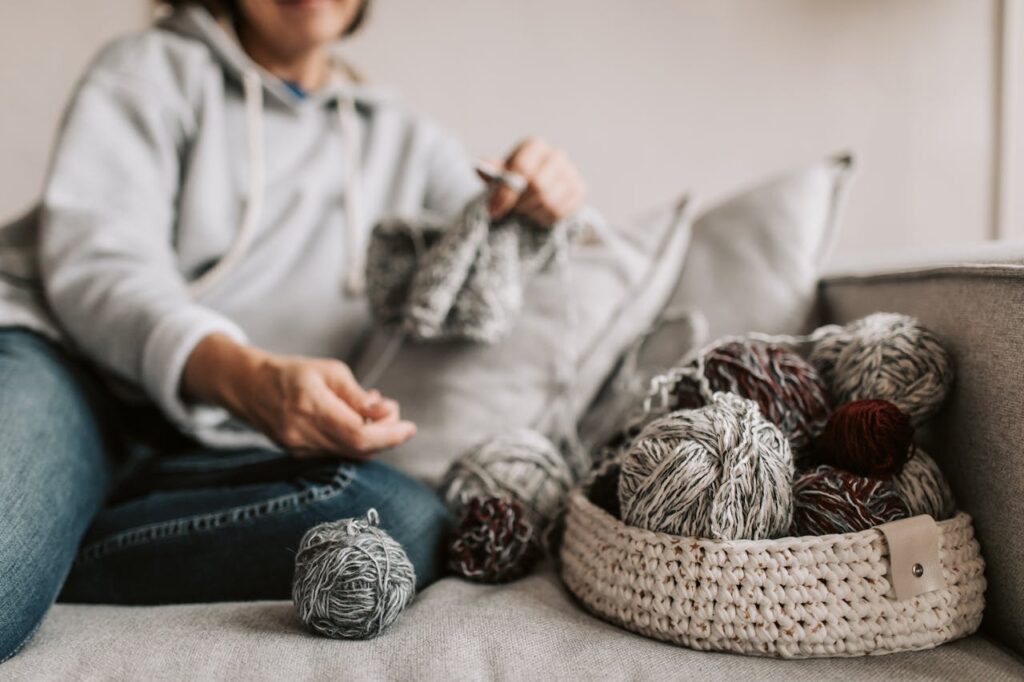 Image of a person sitting on a couch wearing a cozy hoodie, knitting with gray yarn. A woven basket next to them holds multiple skeins of gray and maroon yarn, with a ball of yarn on the couch in the foreground. The scene has a warm and relaxed atmosphere. Representing Knitting or Crocheting.