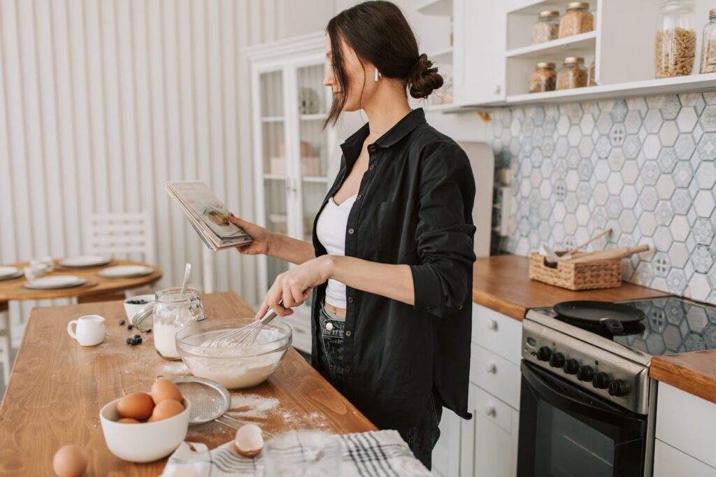 Image of a woman in a kitchen holding a cookbook and whisking ingredients in a glass bowl. The countertop is scattered with flour, eggs, and other baking supplies. The kitchen has a warm, rustic style with hexagonal tile backsplash, wooden countertops, and shelves holding jars of ingredients. A table in the background is set with plates and cups showing  indoor hobbies for women.