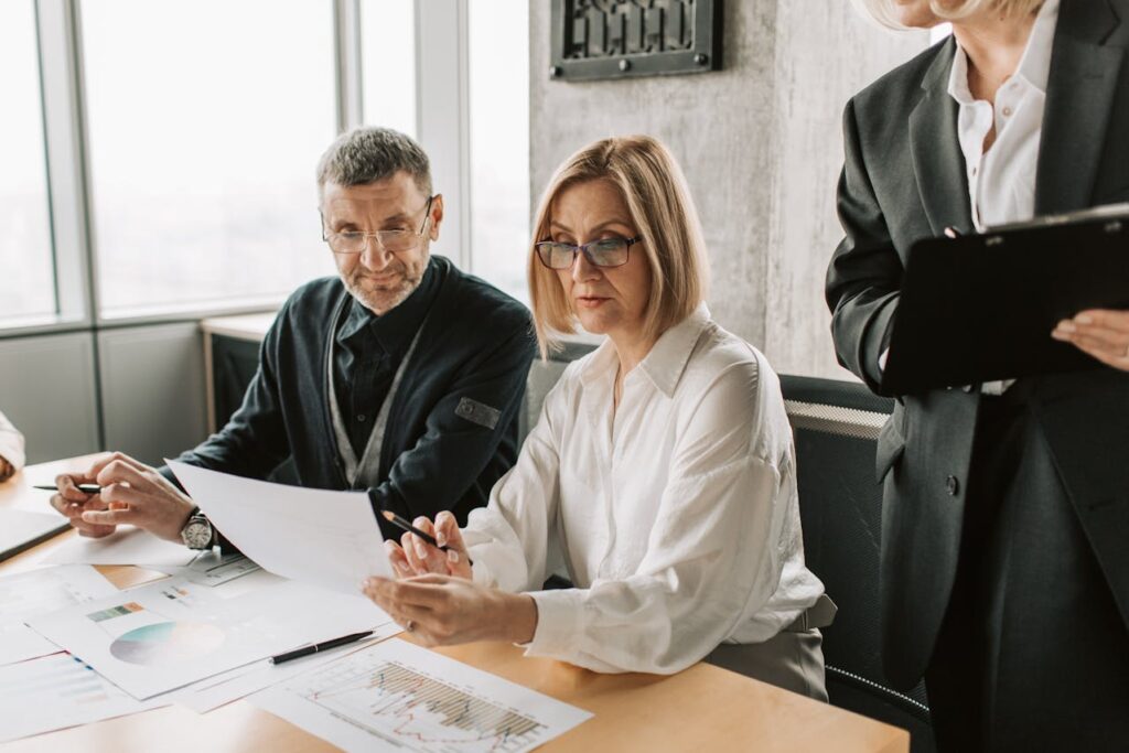 Image of a professional meeting setting with two people seated at a table reviewing documents. A woman in glasses, dressed in a white blouse, is carefully examining a paper while a man next to her, in a dark outfit, looks on attentively. Another individual standing nearby, wearing a suit, is holding a clipboard, observing the discussion. Various charts and documents are spread across the table, indicating a business or data-driven conversation, showing what to wear for the presentation.