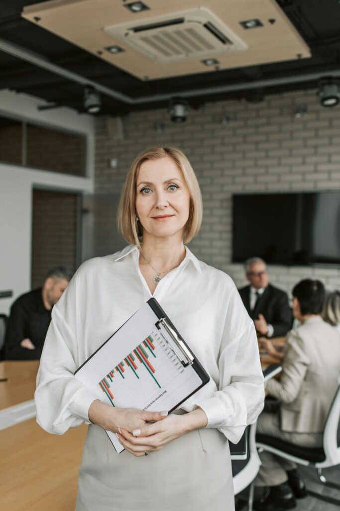 Close-up image of a professional woman is standing in the foreground of a meeting room, holding a clipboard with a bar chart report. She has a calm, confident expression, wearing a white blouse and neutral-colored skirt. The background shows a group of people seated at a conference table, engaged in a discussion, but slightly blurred to focus attention on the woman. The room features a modern design with brick walls and a ceiling-mounted air conditioning unit.