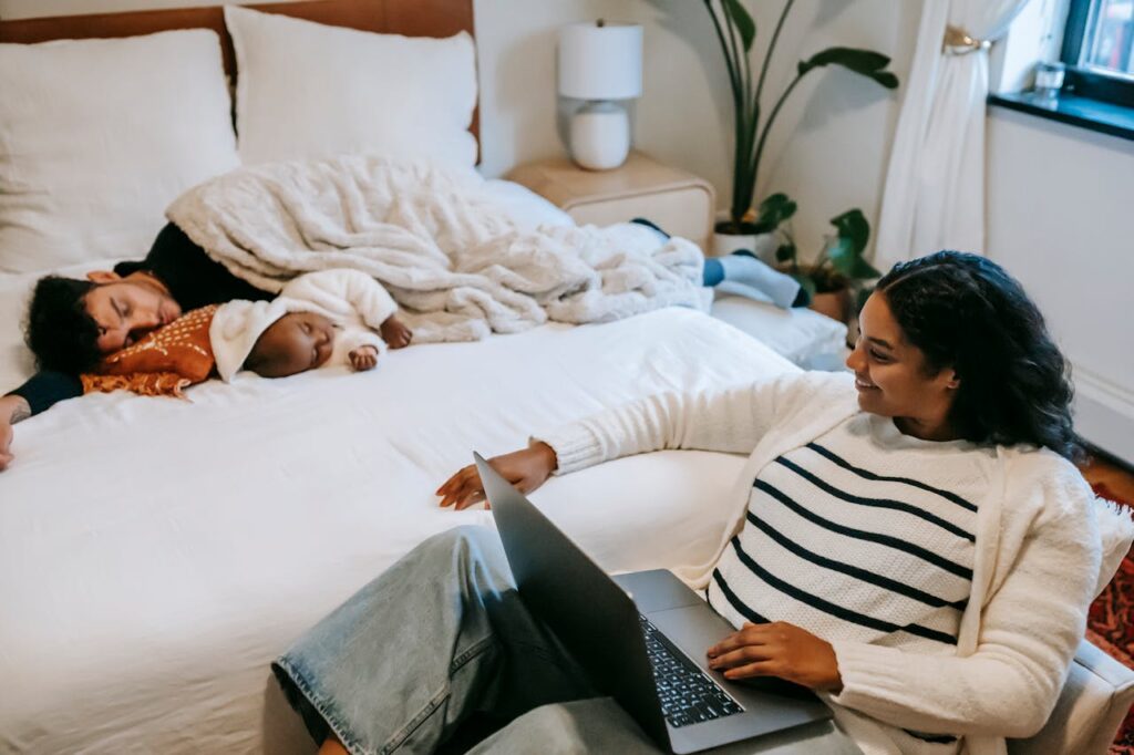 Close-up image of a family scene in a bedroom. A man and a baby are asleep on a bed, cuddled under a soft white blanket. The man is lying on his side with his arm around the baby, who is wearing a light-colored hat and a warm outfit. Next to the bed, a woman sits in a chair, smiling as she looks at them while working on her laptop. She is wearing a striped sweater and light blue jeans, creating a relaxed and comfortable vibe. The room has soft natural lighting, with a plant and a small bedside table in the background, adding to the serene atmosphere.