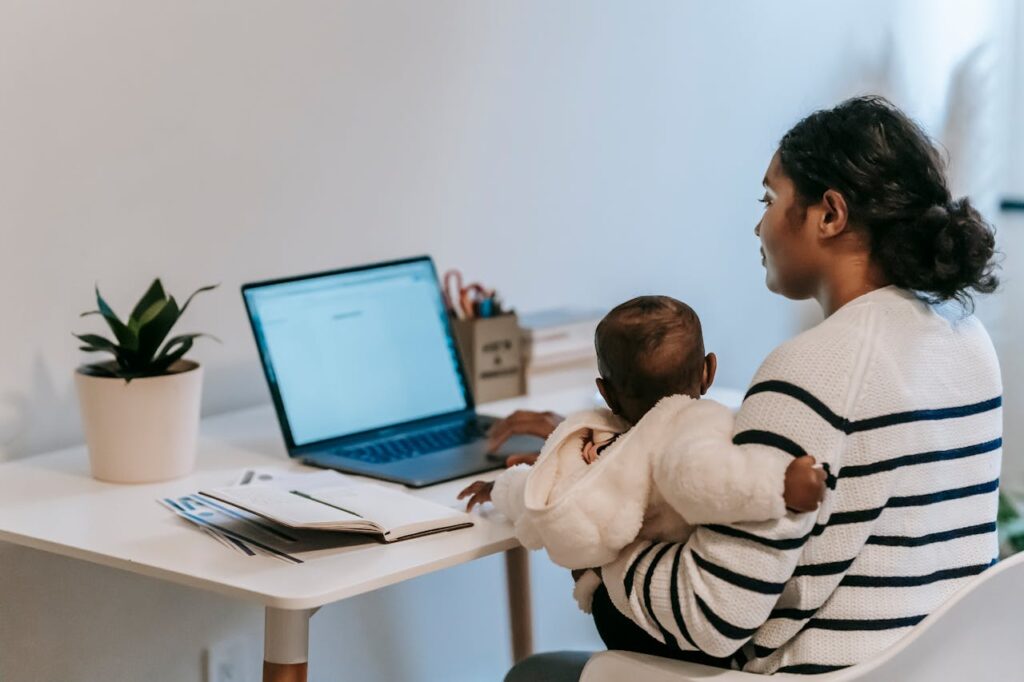 Image of a woman seated at a desk, working on a laptop while holding a baby. The individual is wearing a striped sweater, and the baby is dressed in a cozy white outfit. On the desk, there is a notebook, some papers, and a plant in a pot. The scene suggests a work-from-home environment, balancing professional tasks and childcare. Showing daily routine for working moms