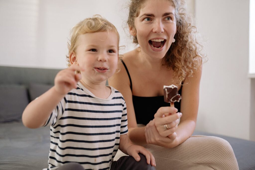 Image of a woman and a child are sitting together, smiling and eating ice cream bars. The child, wearing a striped shirt, looks happy and is holding a small piece of ice cream, while the woman, with curly hair and wearing a black tank top, has an open, joyful expression, holding her ice cream. They appear to be having fun together in a casual and relaxed environment. 