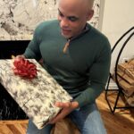 Image of a man sitting on a wooden stool near a fireplace, holding a gift wrapped in festive paper with a red bow, wearing a green sweater and jeans. Image used for the article What to Get Husband for Christmas.