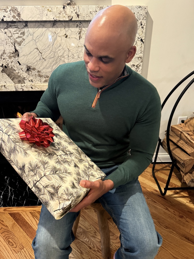 Image of a man sitting on a wooden stool near a fireplace, holding a gift wrapped in festive paper with a red bow, wearing a green sweater and jeans. Image used for the article What to Get Husband for Christmas.