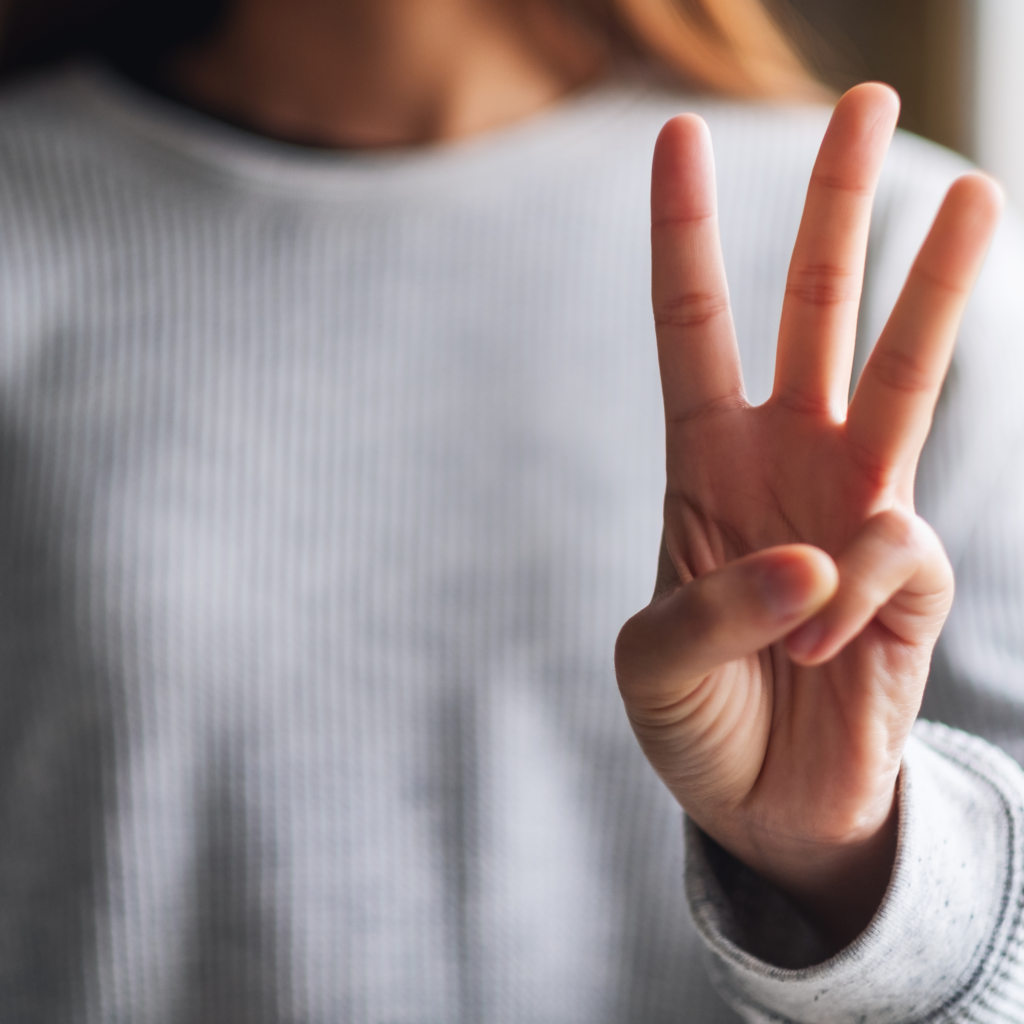 A close-up of a person holding up three fingers, with their hand facing forward. The person is wearing a gray ribbed sweater, and the background is blurred. Representing how to stop cleaning so much.