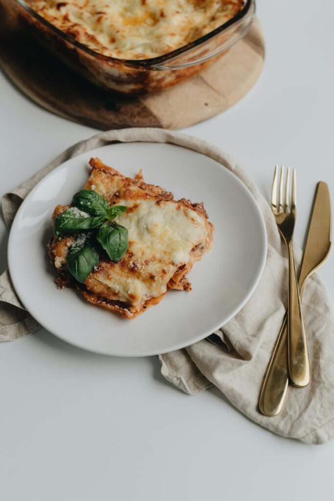 Image of a slice of cheesy lasagna is served on a white plate, garnished with fresh basil leaves. The plate is placed on a light cloth napkin, alongside golden cutlery. A glass baking dish with the remaining lasagna is visible in the background on a wooden board shows working mom cookbook.