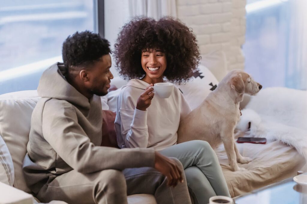 Close-up image of a couple sits on a cozy couch, sharing a joyful moment. The woman, with curly hair and wearing a light hoodie, is holding a cup and smiling warmly at the man beside her. The man, dressed in a casual hoodie, is looking at her with a smile. A dog, possibly a Labrador, sits next to the woman on the couch, looking out towards the window. 