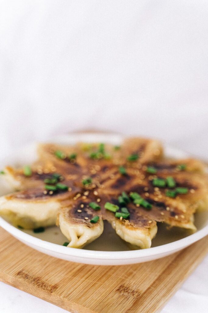 Image of a plate of pan-fried dumplings arranged in a circular pattern, topped with sesame seeds and chopped green onions. The dish is placed on a wooden board with a soft white background showing easy weeknight meals for busy families.