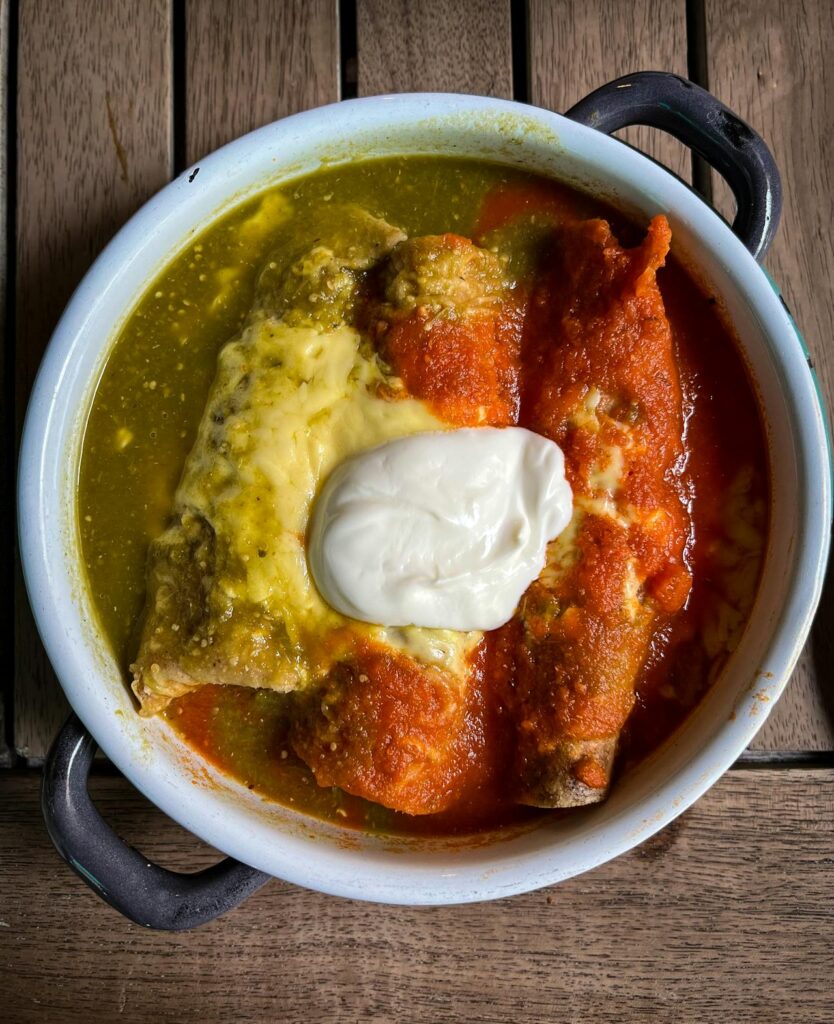 Image of a dish of enchiladas served in a round bowl with handles. The enchiladas are topped with both green and red sauces, melted cheese, and a dollop of sour cream at the center. The dish rests on a wooden surface, representing quick recipes for busy moms.