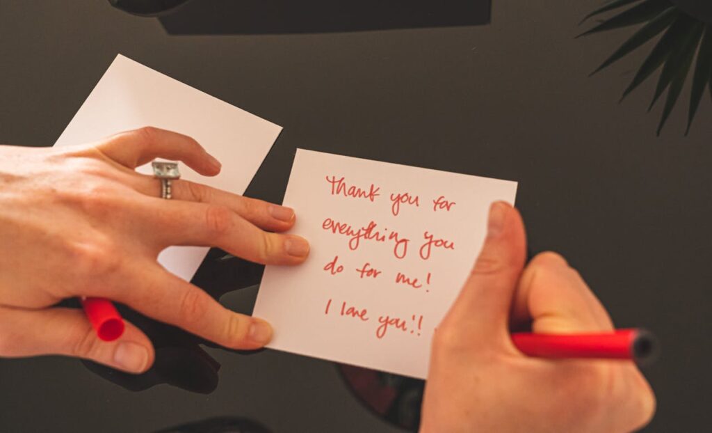 A close-up image of someone writing a heartfelt note on a small piece of paper. The message, written in red ink, says, Thank you for everything you do for me! I love you!! The person’s hands are visible, one holding the paper in place and the other holding a red pen. Another blank card is placed nearby on a dark surface. The atmosphere is warm and personal.