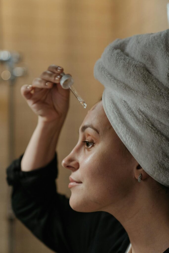 Image of a woman with a towel wrapped around her head is applying a drop of serum or oil to her forehead using a dropper. She has a calm expression and is wearing small hoop earrings and a dark top. The background is softly blurred, with warm lighting.