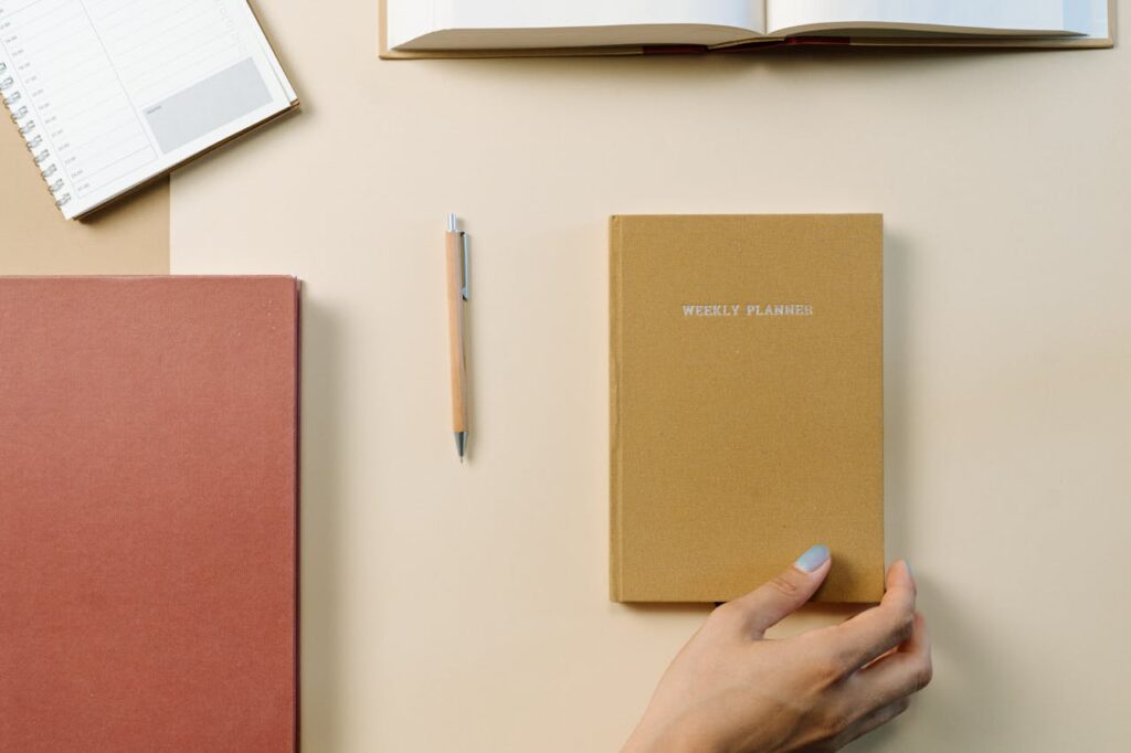 Image of a neatly arranged desk setup featuring a beige weekly planner held by a hand with light blue nail polish, a closed red notebook, a pen, an open book, and a spiral-bound notebook. The items are placed on a beige surface, creating a minimalist and organized aesthetic showing best cleaning hacks for busy people.