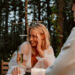 Image of a woman with blonde hair is sitting outdoors at a candlelit dinner table, smiling and holding a glass of champagne. She is wearing a sheer, long-sleeved white blouse and appears to be in a relaxed, joyful moment. The setting is surrounded by tall trees, creating a cozy and intimate atmosphere in a forest. Image showing How Do You Express Gratitude To Your Family.