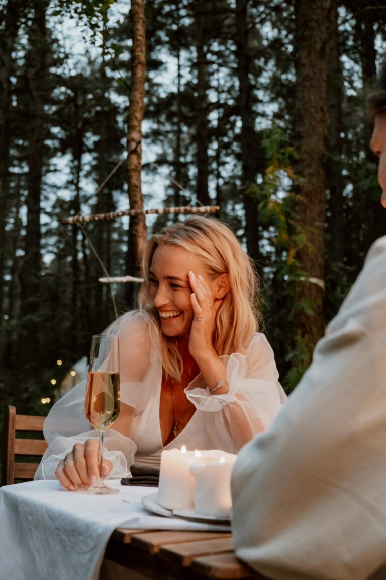 Image of a woman with blonde hair is sitting outdoors at a candlelit dinner table, smiling and holding a glass of champagne. She is wearing a sheer, long-sleeved white blouse and appears to be in a relaxed, joyful moment. The setting is surrounded by tall trees, creating a cozy and intimate atmosphere in a forest. Image showing How Do You Express Gratitude To Your Family.