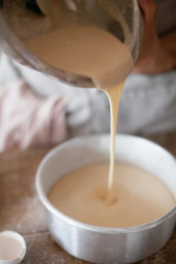 A close-up view of batter being poured from a mixing bowl into a round metal cake pan. A cracked egg shell is visible on the wooden surface nearby, suggesting a baking preparation scene.