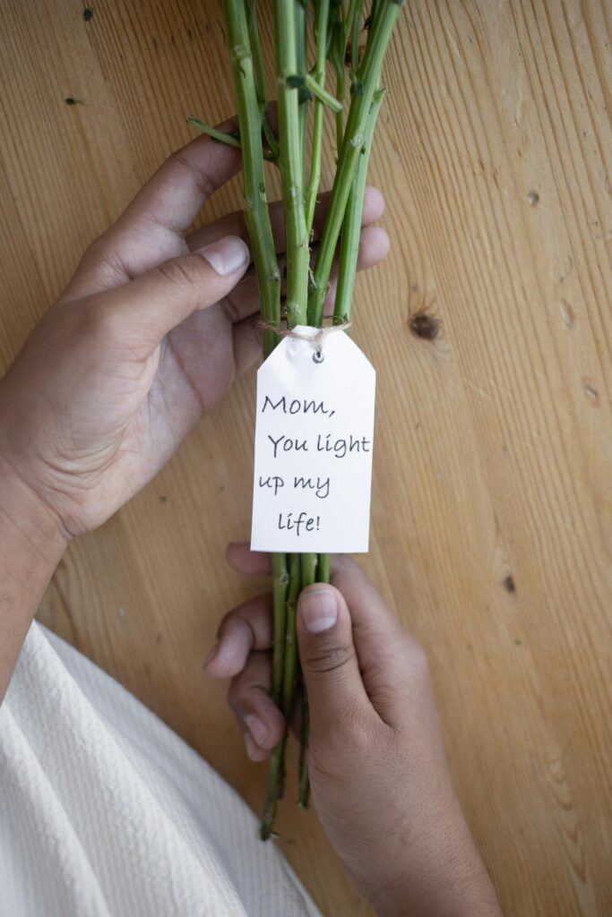 The image shows a pair of hands holding a small bundle of stems tied together, with a white tag attached that reads, "Mom, You light up my life!" The background is a wooden surface. The overall composition suggests a heartfelt, handmade gift or gesture for a mother.