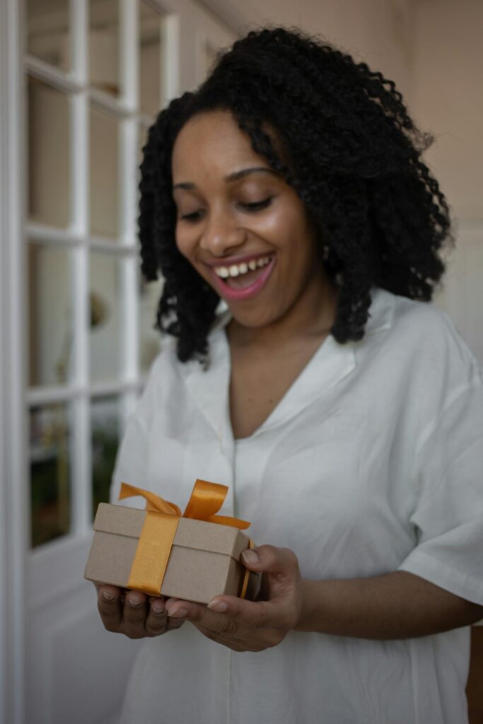 Close-up image of a woman with curly hair and a joyful expression is holding a small gift box wrapped with a yellow ribbon. She is dressed in a white shirt and is standing indoors, looking at the gift with excitement. The scene captures a moment of surprise and happiness representing gratitude for family.